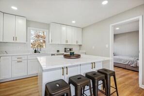 A modern kitchen with white cabinets, a large island with stools, and a view to a bedroom.