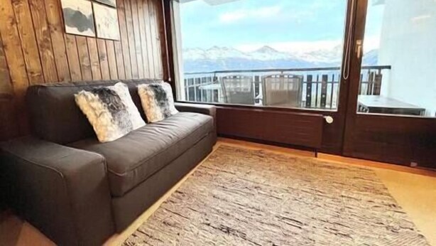 A room with a brown leather sofa, a large window with a view of mountains, and a wooden floor. - Cosy studio for 5 near ski lift, Les Gentianes (Les Collons)