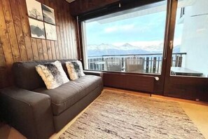 A room with a brown leather sofa, a large window with a view of mountains, and a wooden floor. - Cosy studio for 5 near ski lift, Les Gentianes (Les Collons)