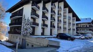 A multi-story building with balconies, a snow-covered ground, and parked cars. - Cosy studio for 5 near ski lift, Les Gentianes (Les Collons)