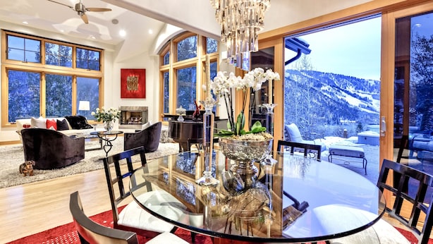 Ein modernes Wohnzimmer mit einem Glastisch, schwarzen Stühlen und einer Chandelier. Große Fenster bieten Blick auf eine verschneite Landschaft.