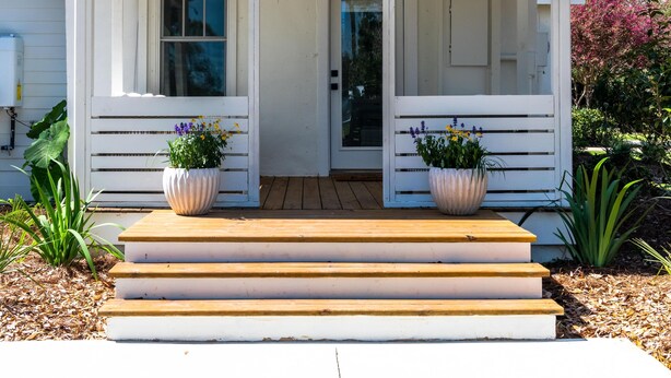 A white single-story house with a covered porch, two large potted plants, and a wooden deck leading to the entrance. - Palmetto Bungalow - Quaint, original Fairhope cottage close to town. (Fairhope)
