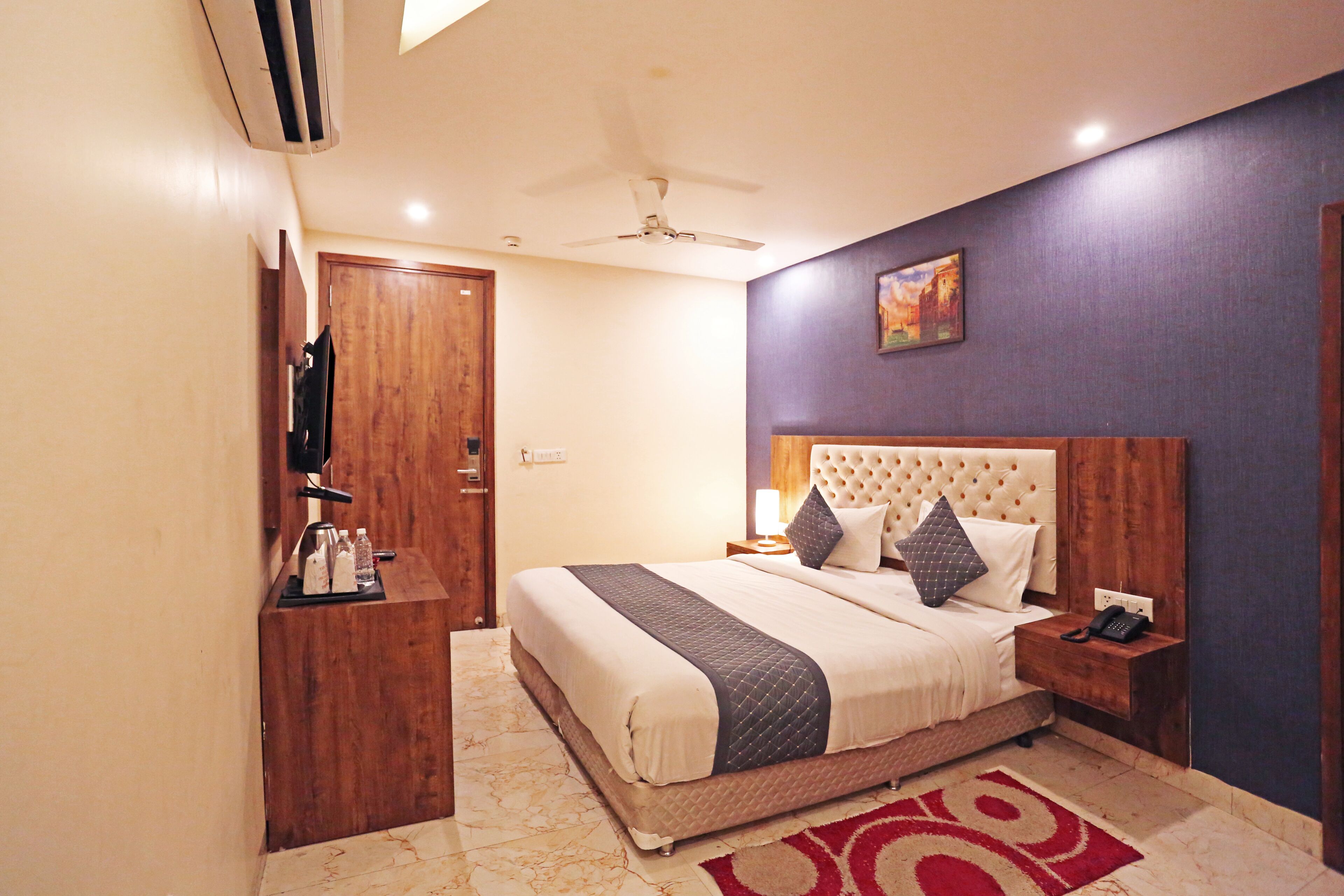 A hotel room with a bed, a wooden nightstand, a telephone, a wall-mounted TV, and a ceiling fan.