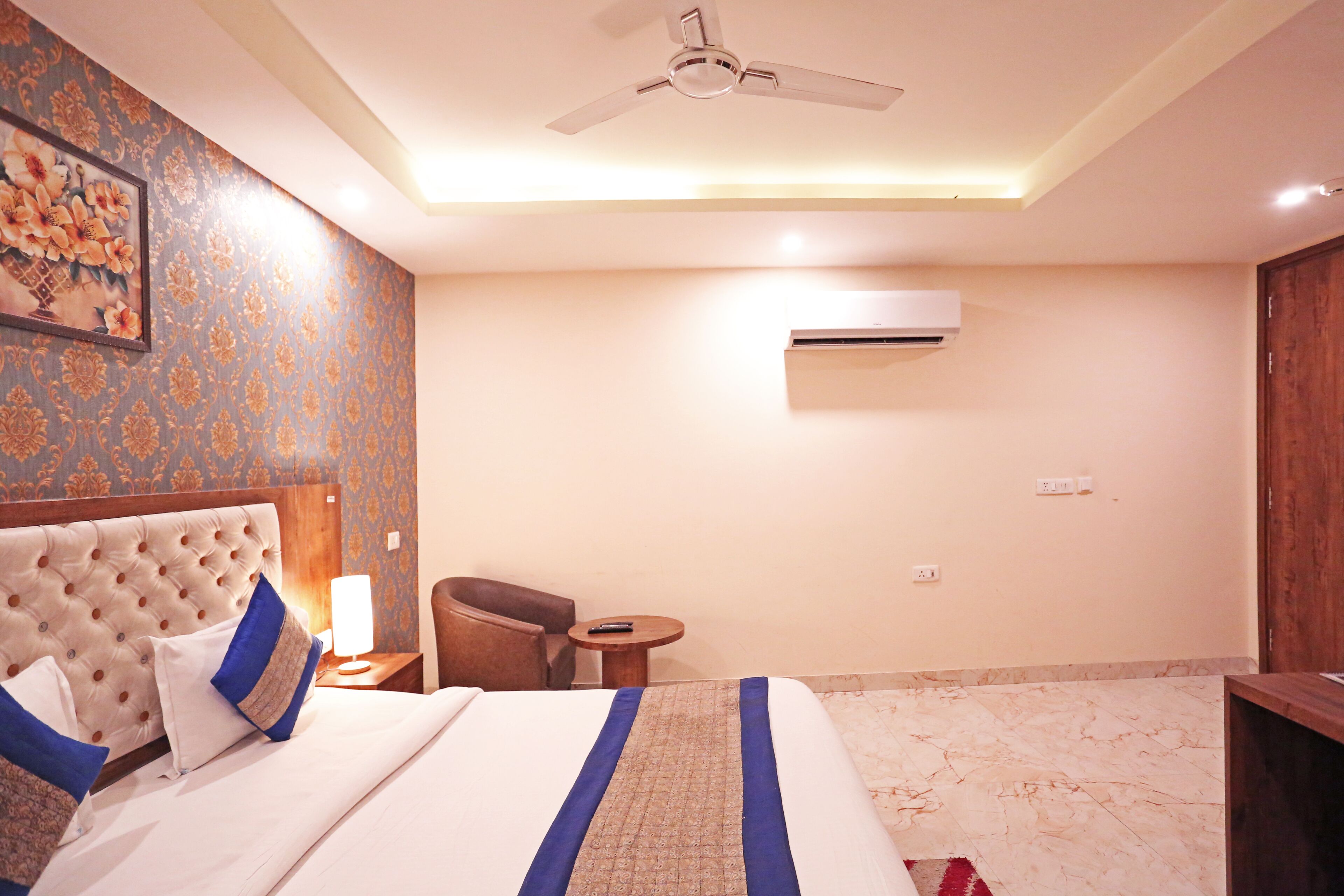 A hotel room with a tufted headboard, a bed with white and blue bedding, a small round table with a lamp, a chair, a ceiling fan, and an air conditioning unit on the wall.