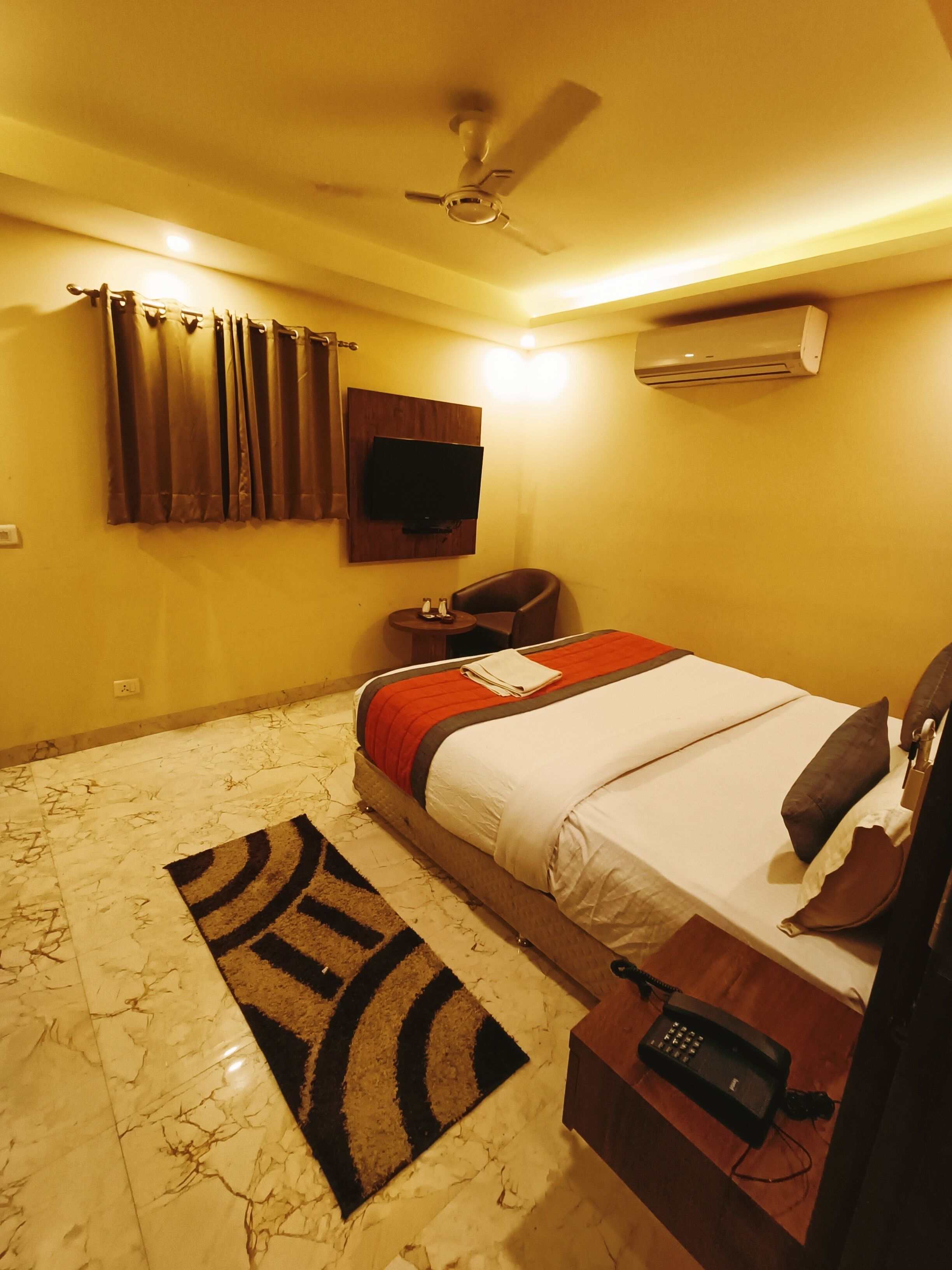 A hotel room with a bed, a desk with a telephone, a chair, a television mounted on the wall, an air conditioner, and a ceiling fan.