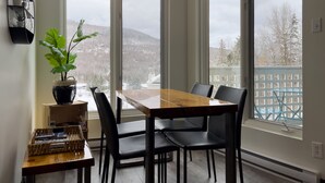 A dining area with a wooden table and black chairs, large windows with a view of snow-covered hills, and a potted plant on the left. - Loft Ski-in/Ski-out Stoneham resort (Stoneham-et-Tewkesbury)