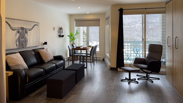 A modern living room with a black leather sofa, a dining table with chairs, a black office chair, and a large window with a view of a snowy landscape. - Loft Ski-in/Ski-out Stoneham resort (Stoneham-et-Tewkesbury)