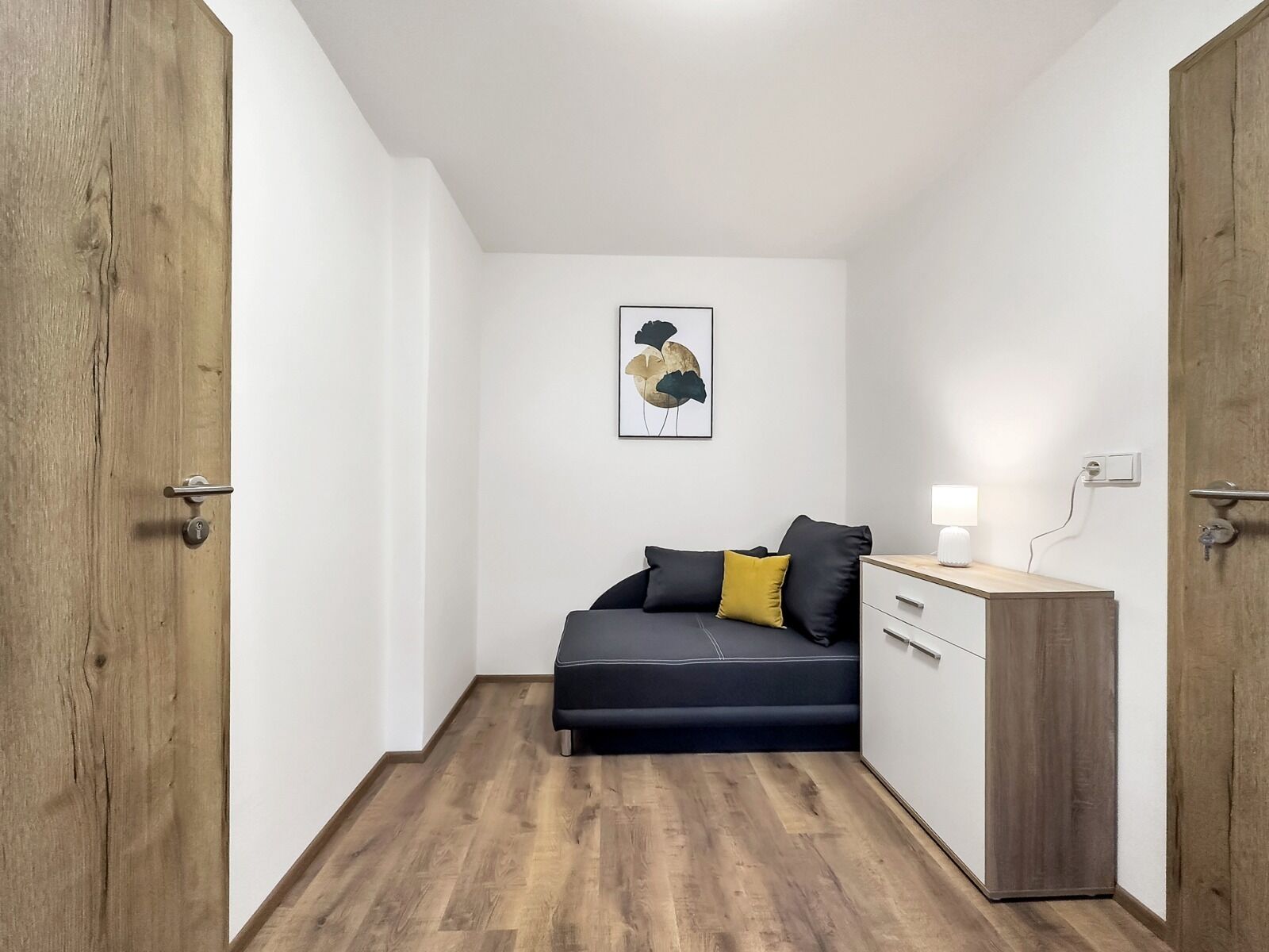 Una habitación pequeña y moderna con suelo de madera, un sofá oscuro con cojines, un escritorio pequeño con lámpara y un cuadro en la pared.