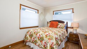 Un dormitorio con cubrecama floral, cabecera de madera, mesita de noche con lámpara y dos ventanas con persianas.