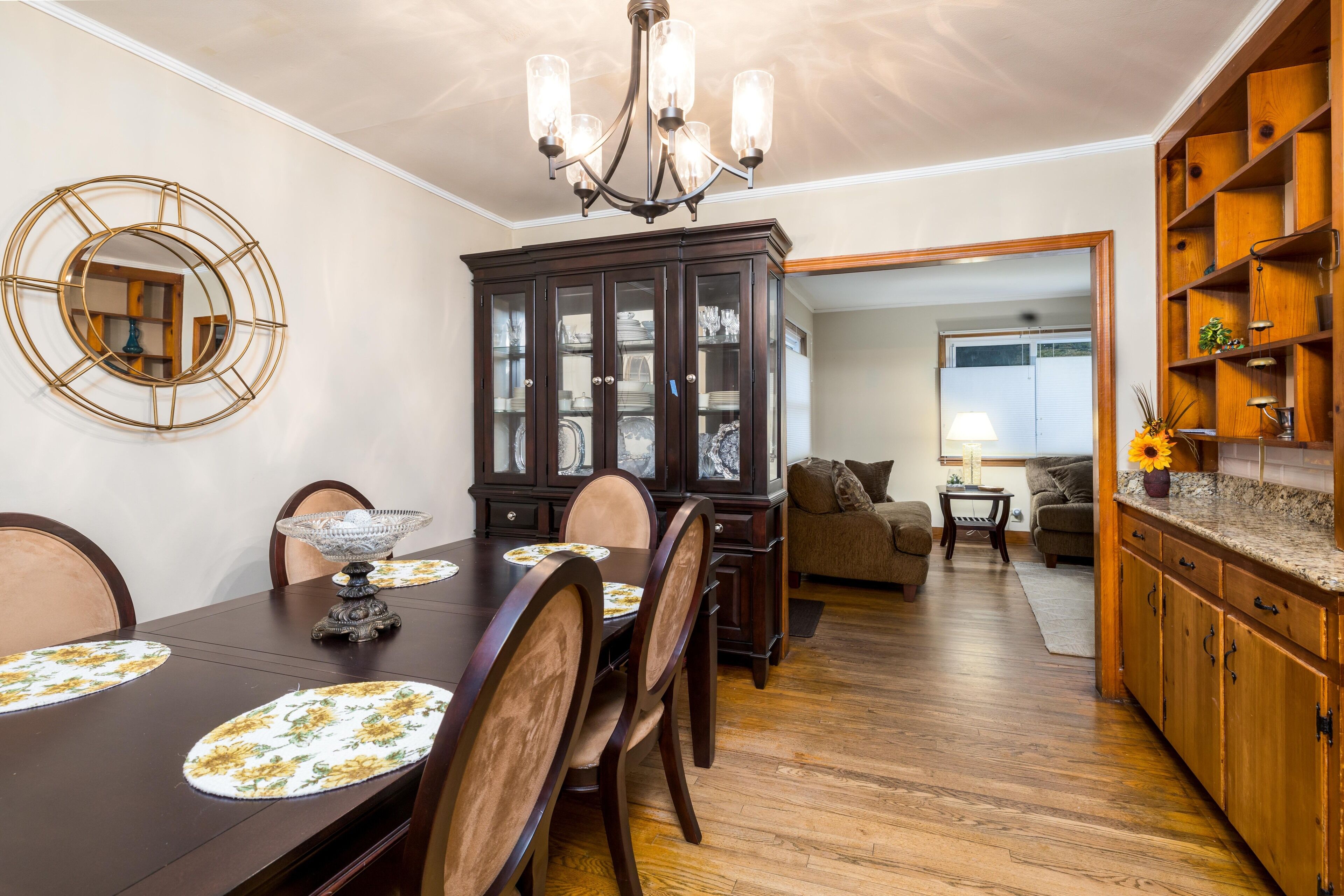A dining area with a dark wooden table, chairs, and a chandelier. There is a cabinet with glass doors and a mirror on the wall.