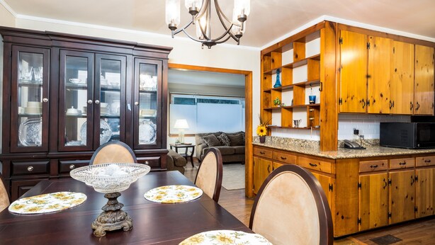 Comedor con mesa de madera, sillas y una araña. Zona de cocina con armarios de madera y un microondas.