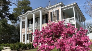 A large, two-story house with a porch and columns, surrounded by a well-maintained lawn and a flowering tree.