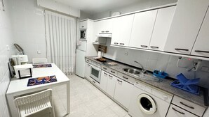 Cocina compacta con armarios blancos, horno integrado, lavadora y un pequeño comedor con una silla.