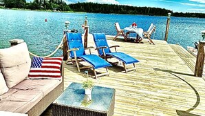 A wooden deck with lounge chairs and a sofa, overlooking a calm lake with a forested shoreline. - Log house with private beach and own dock incl. boat (Njurunda)