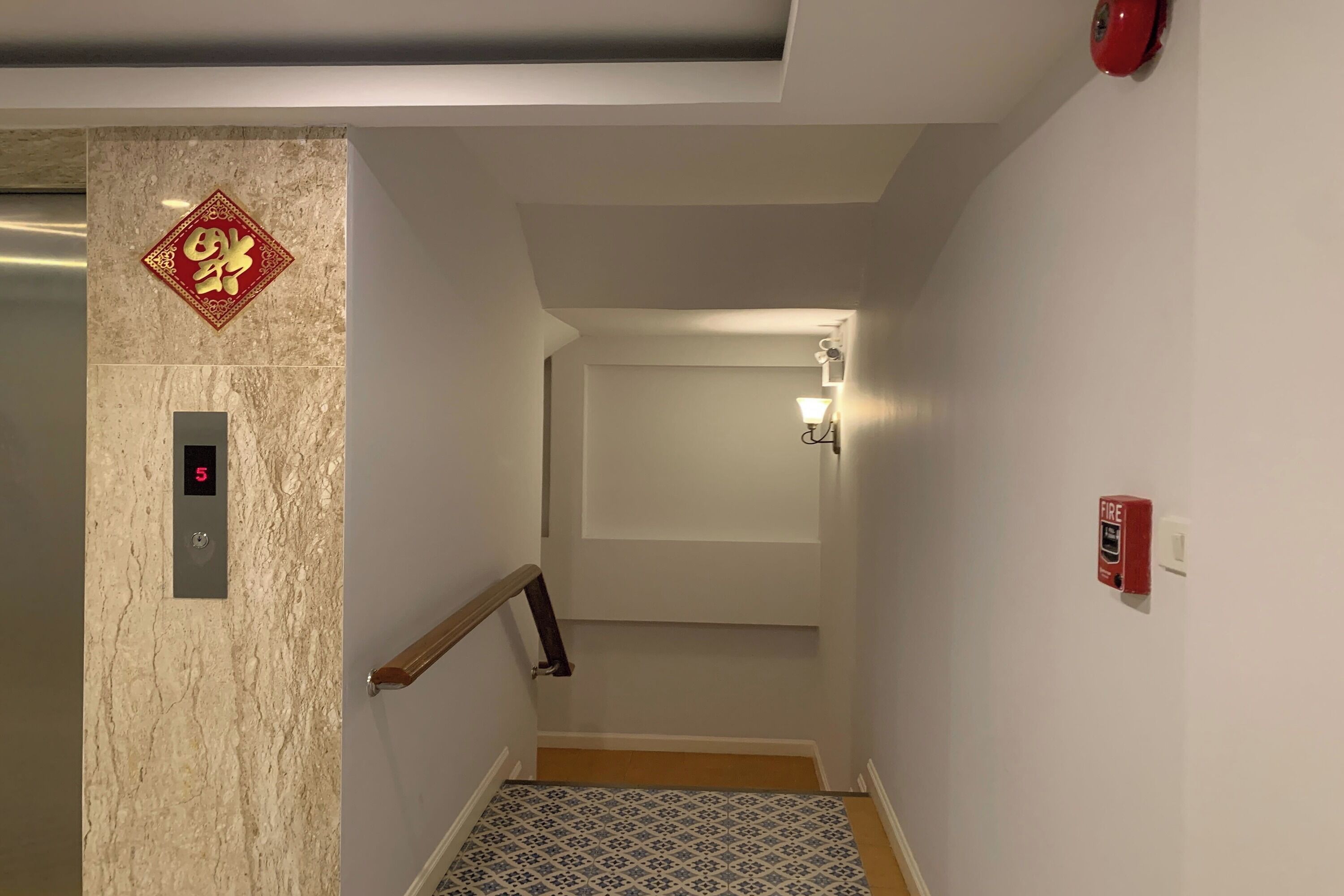 A narrow hallway with a marble wall, a fire alarm, and a door at the end.