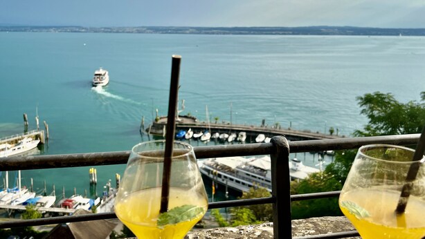 A view of a marina with boats docked, a ferry in the water, and two glasses of yellow drink on a railing.