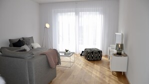 A modern bedroom with a grey sofa, a bedside table with a lamp, a small white cabinet, and a window with sheer curtains.