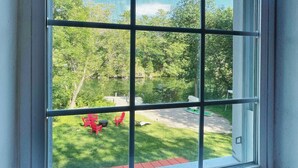 A window with a view of a green lawn and a red chair. - Harmonious Cottage by the river (Washago)