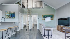Un espacio habitable estilo loft con cama en zona alta, una escalera, comedor con taburetes, sofá, televisor y obras de arte en las paredes.