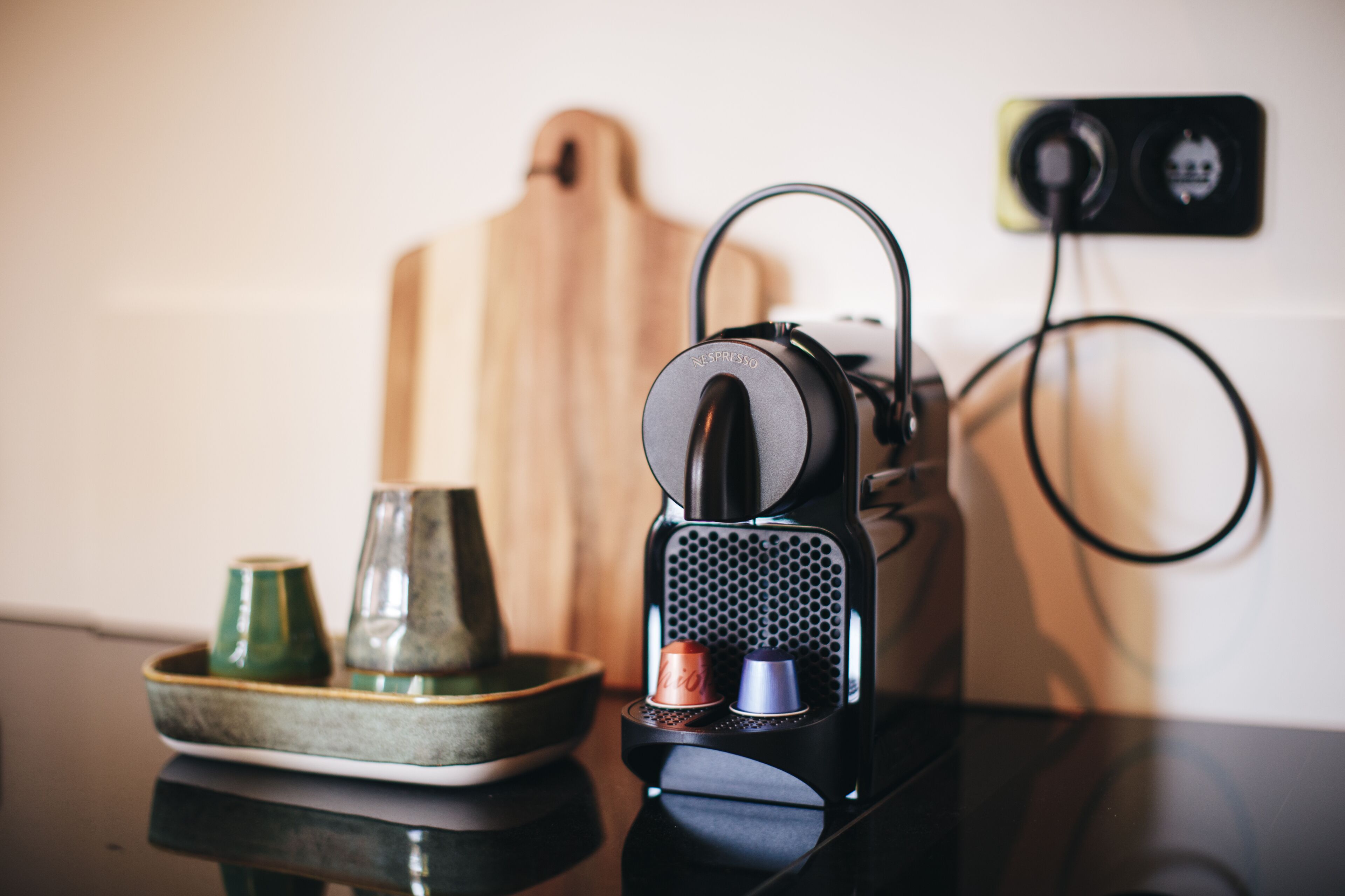 A coffee machine with a cup and a glass on a tray, a power outlet, and a wooden board in the background.