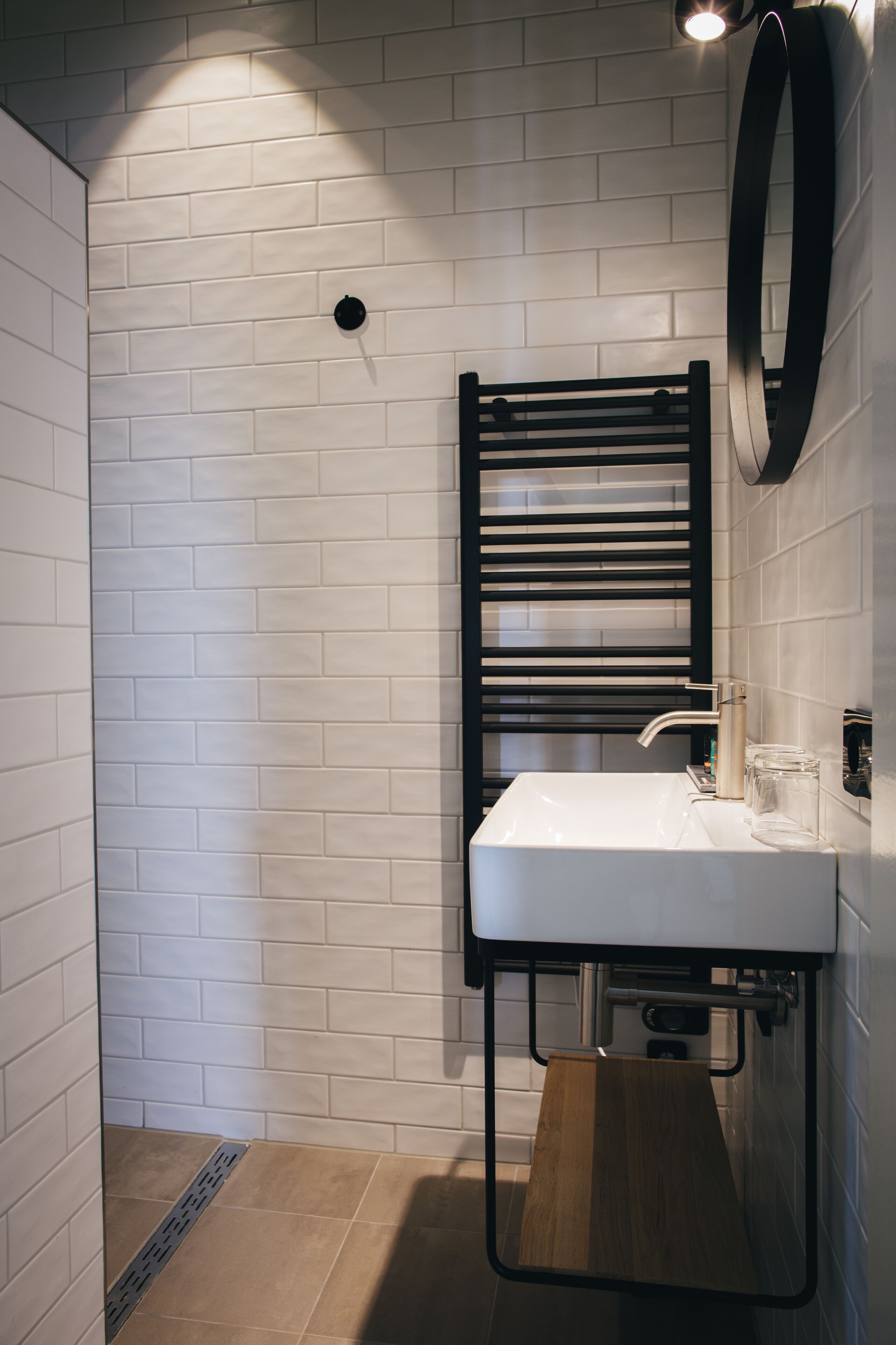 Une salle de bain avec un mur recouvert de carreaux blancs, un porte-serviettes noir, un lavabo blanc et un miroir.