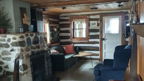 A log cabin interior with a stone fireplace, wooden floors, a sofa, a blue armchair, and a small table. - Waterfront Modernized Rustic Log Cabin on Indian Lake, Manistique, MI (Manistique)