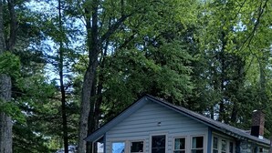 A rustic cabin with a porch, surrounded by a natural landscape with a small stream. - Waterfront Modernized Rustic Log Cabin on Indian Lake, Manistique, MI (Manistique)