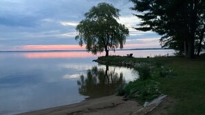 A lakeside scene with a tree, a sandy beach, and a calm water body reflecting the sky. - Waterfront Modernized Rustic Log Cabin on Indian Lake, Manistique, MI (Manistique)