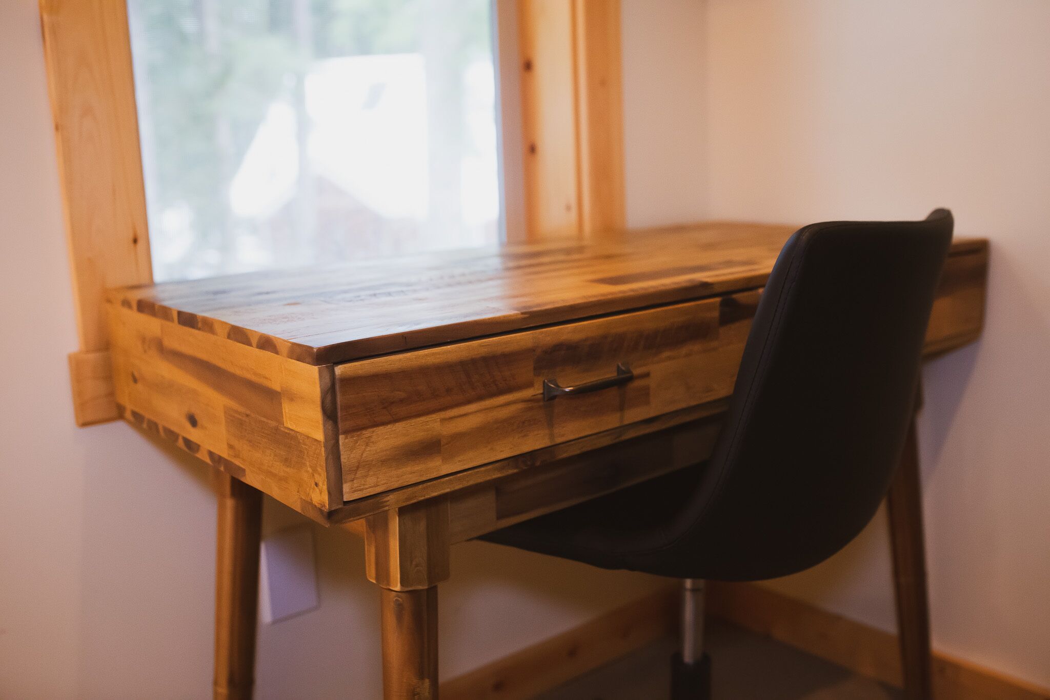 A wooden desk with a black chair, a drawer, and a window in the background.