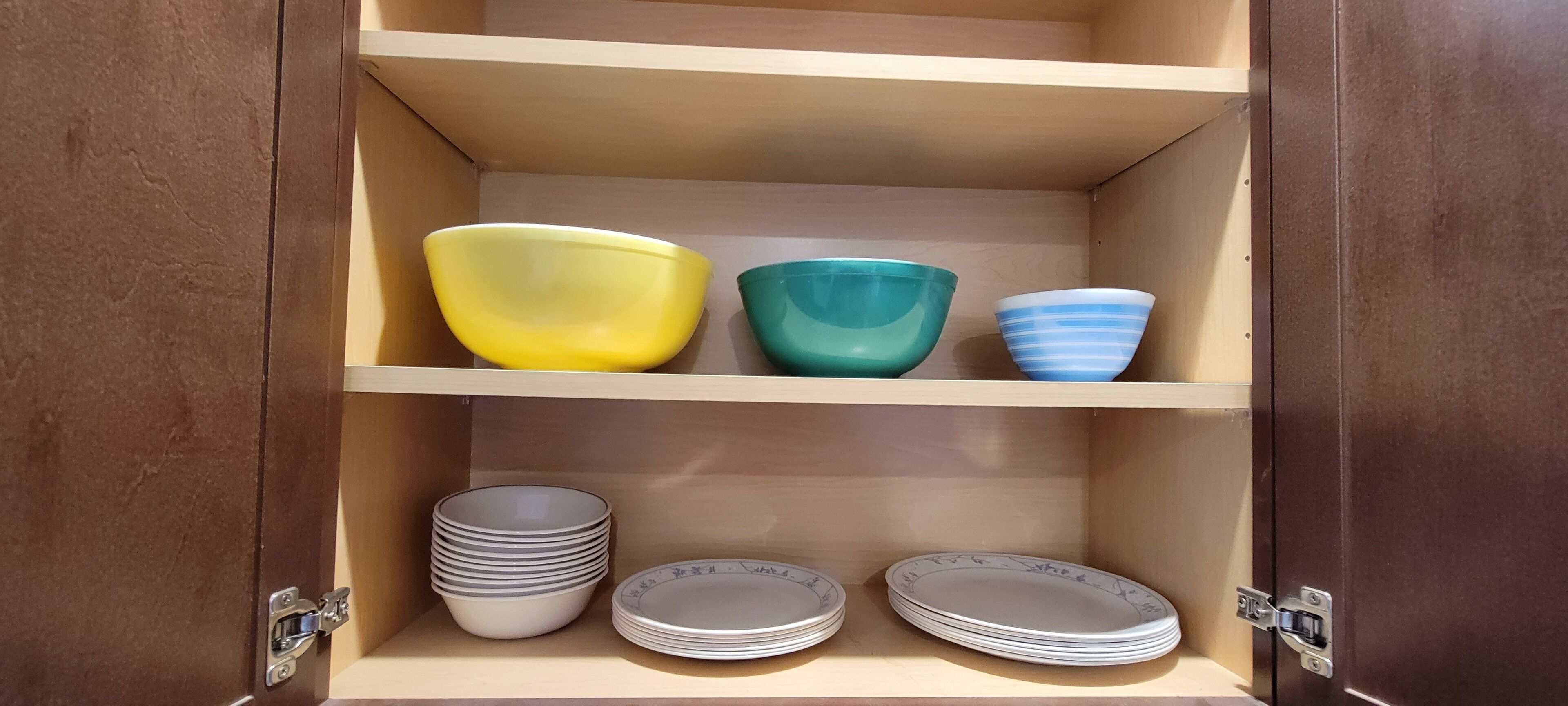 A wooden cabinet with three shelves. The top shelf holds three bowls in yellow, green, and blue. The middle shelf has stacks of white plates with a floral pattern. The bottom shelf also has stacks of white plates with a floral pattern.