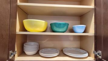 A wooden cabinet with three shelves. The top shelf holds three bowls in yellow, green, and blue. The middle shelf has stacks of white plates with a floral pattern. The bottom shelf also has stacks of white plates with a floral pattern.