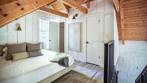 Une chambre avec un grand lit, des poutres apparentes en bois au plafond, une porte blanche et une fenêtre.