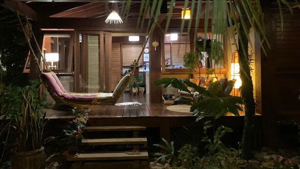 Een houten terras met een hangmat en een bankje, omringd door tropische planten.