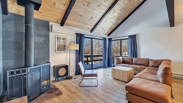 A modern living room with a fireplace, wooden ceiling beams, a sofa, and a large window. - Modern and Brand New, Pool, Camelback Ski Area, 2 showers (Tannersville)