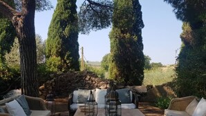 Un espacio al aire libre con mobiliario de mimbre y faroles, rodeado de árboles altos y un muro de piedra.