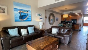 Un vaste salon avec un grand canapé en cuir marron, une table basse en bois et un ventilateur de plafond. Un tableau représentant un bateau est accroché au mur, et on aperçoit une cuisine au fond.