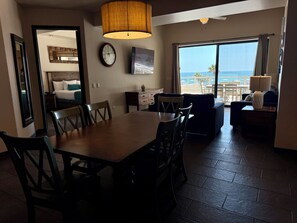 A dining area with a table and chairs, a clock on the wall, and a view of the ocean through large windows.