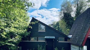 Une maison en bois sombre, avec un toit triangulaire, une porte d’entrée et une grande fenêtre.