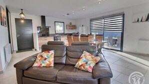 A modern living room with a brown leather sofa, two colorful pillows, a dining area with wicker chairs, and a kitchen in the background. - Villa Armor Azur - ERQUY (ERQUY)