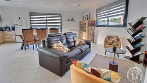 A modern living room with a leather sofa, a dining area with wicker chairs, a bookshelf, and large windows with blinds. - Villa Armor Azur - ERQUY (ERQUY)