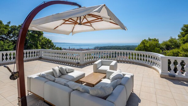 A sunlit outdoor seating area with a white sofa and a wooden table, overlooking a scenic view of the sea and distant landscape. - Beautiful home in Rab (Rab)