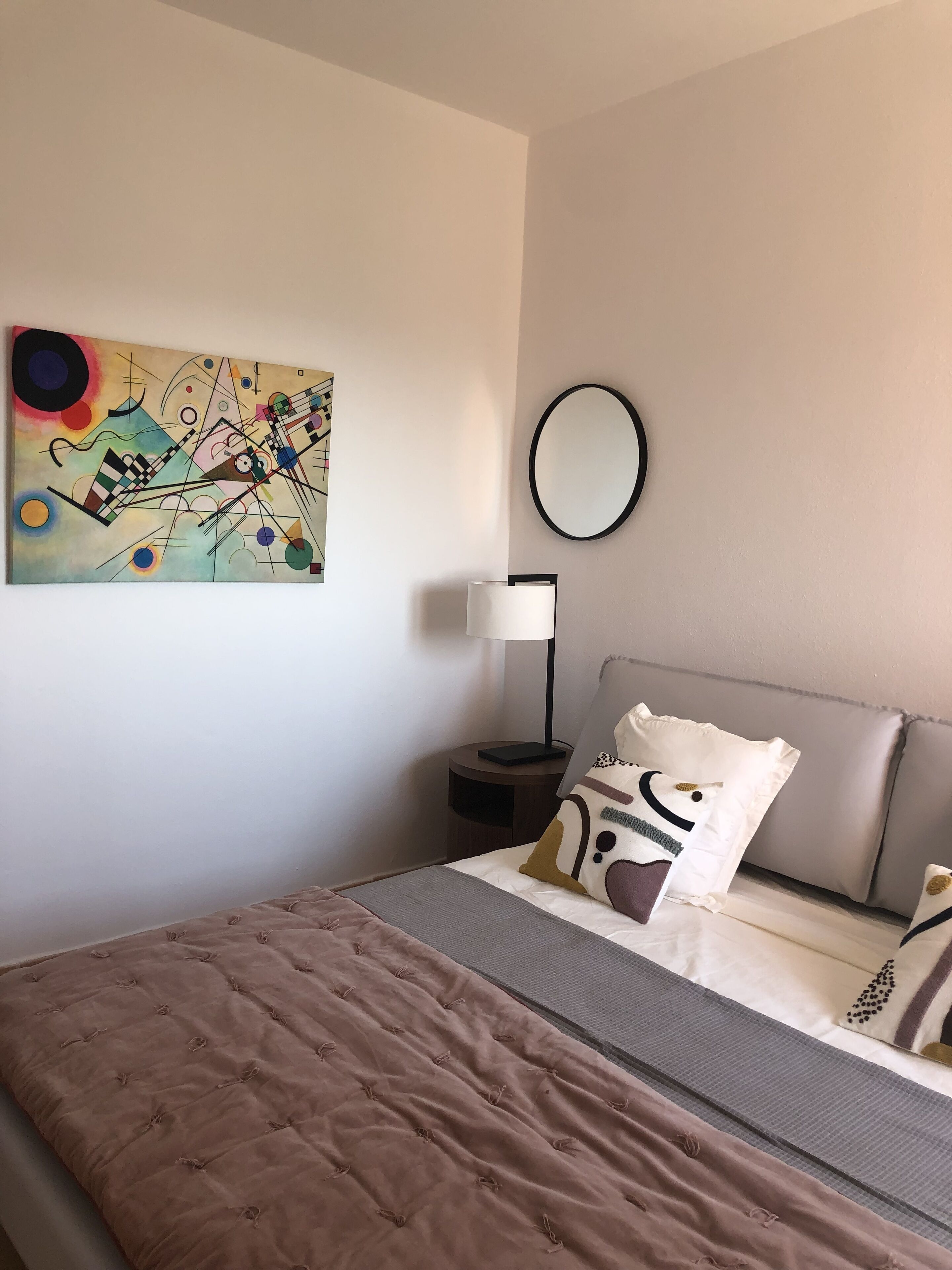 A neatly arranged hotel room with a bed, a bedside table with a lamp, a wall-mounted mirror, and a colorful abstract painting.