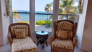 A room with two wicker chairs and a small round table, large windows with a view of the ocean and palm trees.