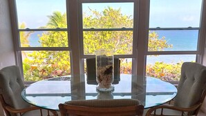 A round glass table with two wicker chairs, a vase with flowers, and a view of the ocean through large windows.
