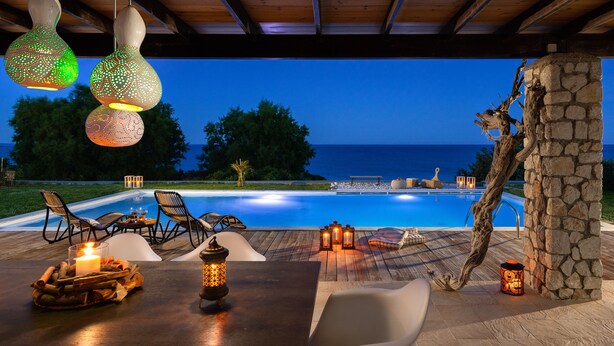 An outdoor pool area with a wooden deck, lounge chairs, and a stone pillar. - FZIN Villa Sevil (Rhodes)