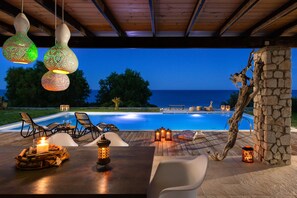 An outdoor pool area with a wooden deck, lounge chairs, and a stone pillar. - FZIN Villa Sevil (Rhodes)