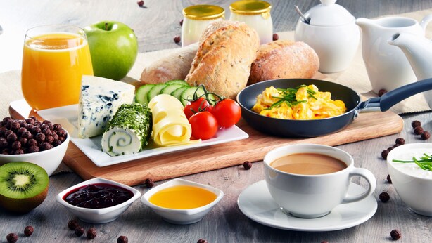 A breakfast setting with various food items, a jug of orange juice, a bowl of cereal, a plate with eggs and vegetables, and a cup of coffee.