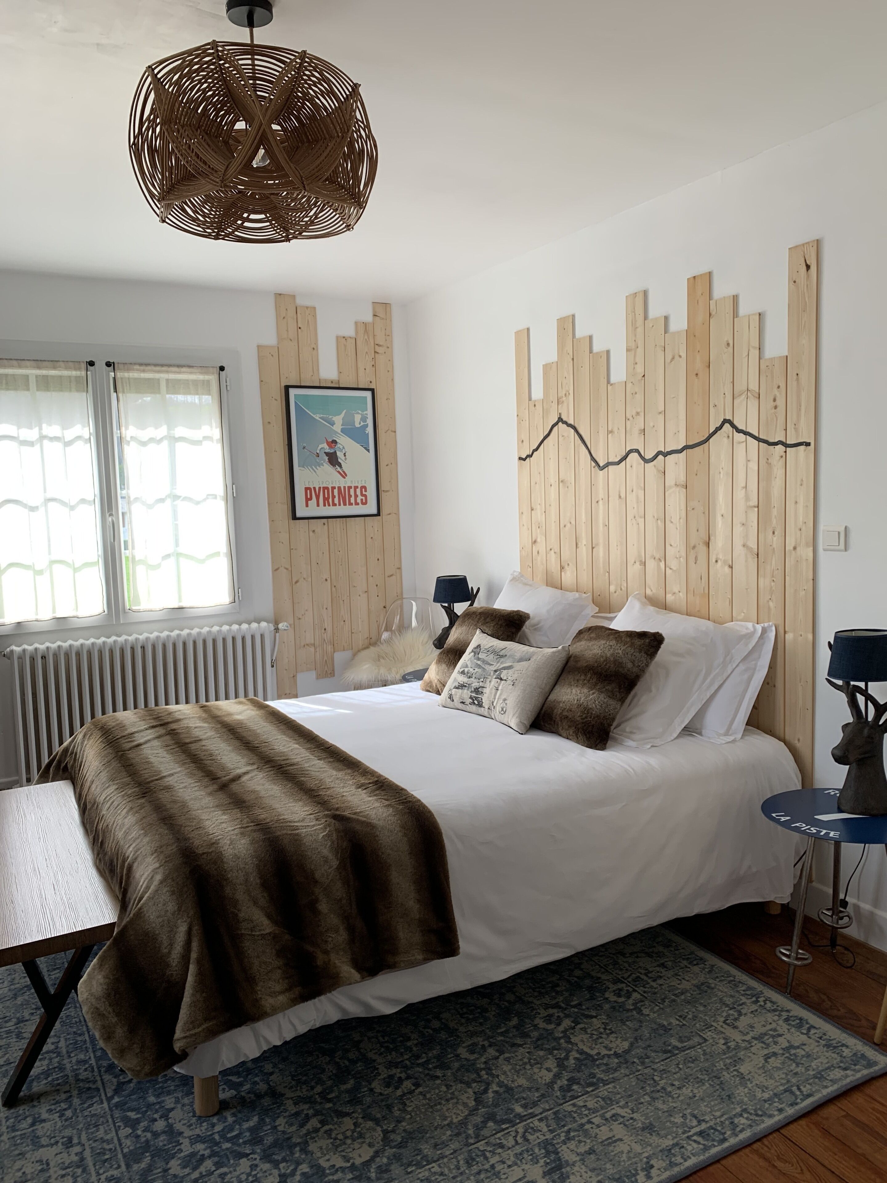 A bedroom with a large bed, a wooden headboard, a poster on the wall, a window with curtains, a radiator, a table lamp, and a decorative wall piece.