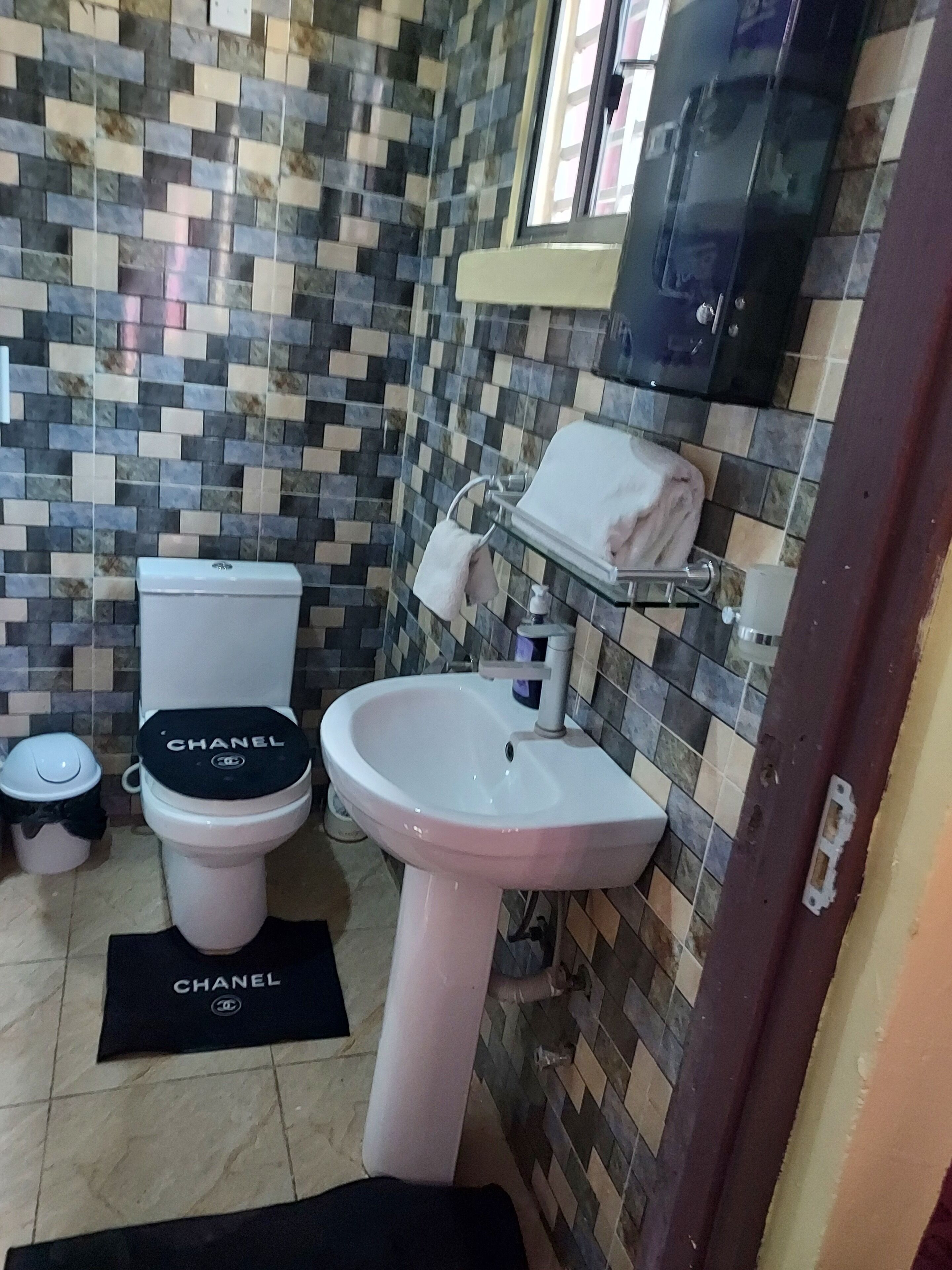 Een badkamer met een betegelde muur, een toilet met een zitje met 'CHANEL'-branding, een wastafel en een handdoekhouder.