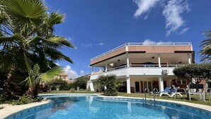 A swimming pool with a clear blue water surface, surrounded by palm trees and a two-story building with a balcony. - Annas Garden (Torrevieja)